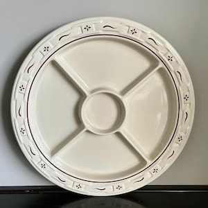 May include: A round, off-white ceramic serving platter with a divided design. The platter features a central circular well and four pie-shaped compartments. A burgundy floral and wavy line pattern decorates the rim. The platter is ideal for serving appetizers or snacks.