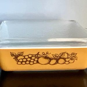 May include: Rectangular, amber-colored Pyrex baking dish with a clear, ribbed glass lid. The dish features a brown fruit design around the sides. A vintage kitchenware item.