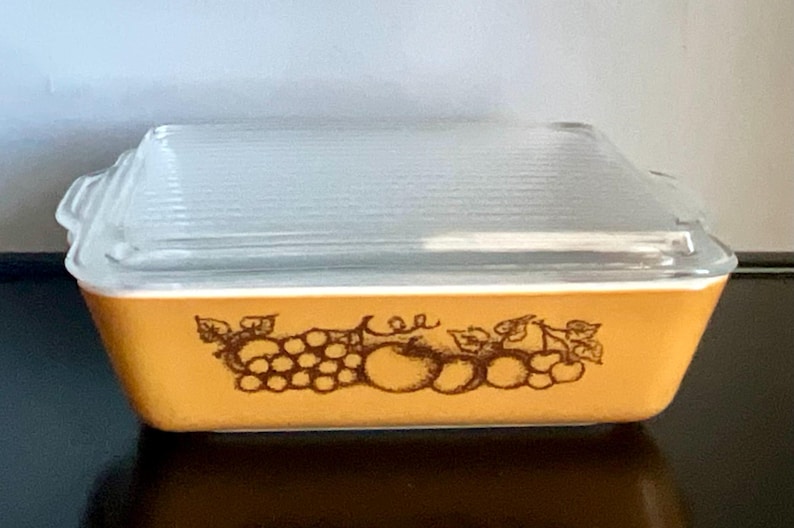 May include: Rectangular, amber-colored Pyrex baking dish with a clear, ribbed glass lid. The dish features a brown fruit design around the sides. A vintage kitchenware item.