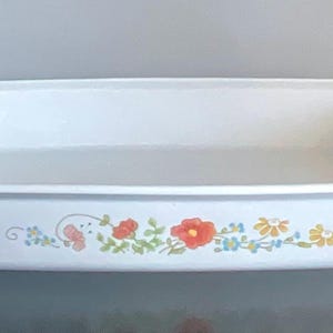 May include: White rectangular baking dish with a floral design. The dish has a slightly raised rim and is decorated with red, orange, yellow, and blue flowers and green leaves. The dish is sitting on a dark surface.