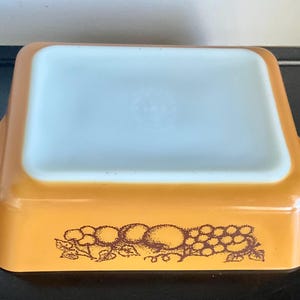 May include: An amber-colored rectangular Pyrex baking dish with a white interior. The dish features a brown fruit design on the exterior. The dish has two handles and is designed for oven use.