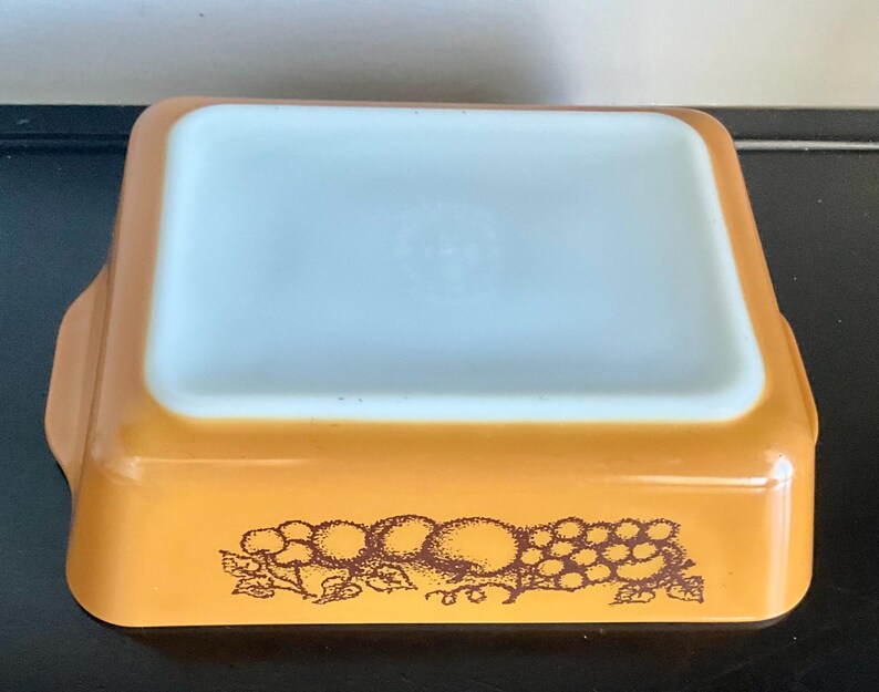May include: An amber-colored rectangular Pyrex baking dish with a white interior. The dish features a brown fruit design on the exterior. The dish has two handles and is designed for oven use.