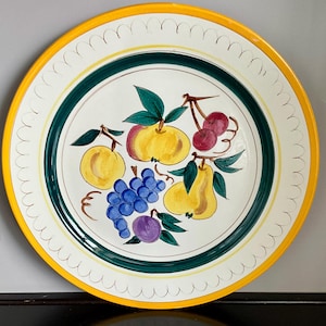 May include: A large, round decorative plate with a yellow rim and a white center. The plate features a hand-painted design of various fruits, including yellow apples, purple grapes, and red cherries, with green leaves. A green and yellow border surrounds the fruit design.