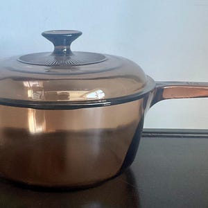May include: A translucent amber-colored glass saucepan with a matching lid. The pot features a single long handle on one side and a small tab handle on the opposite side. The lid has a central knob and a circular textured pattern on its top surface.
