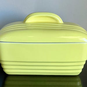 May include: A rectangular, light yellow ceramic butter dish with a matching lid. The lid has a curved handle and the dish features horizontal ridges. The butter dish is sitting on a dark surface.