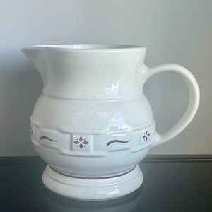 May include: White ceramic pitcher with a rounded body, a spout, and a curved handle. The pitcher features a decorative band with floral and curved accents in a dark red color. The pitcher sits on a circular base.