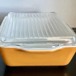 May include: A vintage, square-shaped glass dish with a clear, ribbed lid. The base is a solid, warm orange color, and the lid has a slightly angled design. The dish is likely used for food storage or serving.