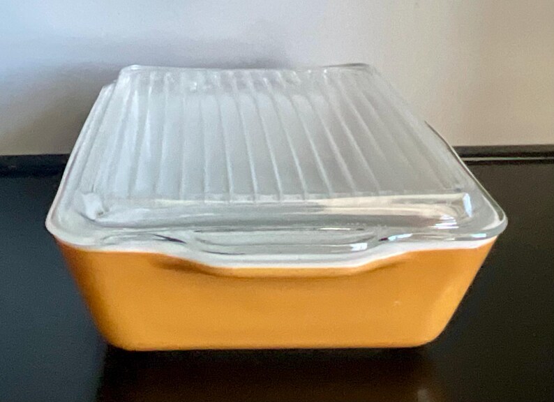 May include: A vintage, square-shaped glass dish with a clear, ribbed lid. The base is a solid, warm orange color, and the lid has a slightly angled design. The dish is likely used for food storage or serving.