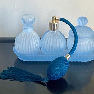 May include: A set of three light blue frosted glass vanity items on a matching rectangular tray. The set includes a bottle with a stopper, a perfume atomizer with a dark teal bulb and tassel, and a lidded jar, all featuring vertical ribbing.