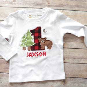 Peut inclure: T-shirt &agrave; manches longues blanc avec un num&eacute;ro un en tissu &eacute;cossais rouge et noir, un arbre vert, un ours brun et le nom Jaxson brod&eacute; sur le devant.