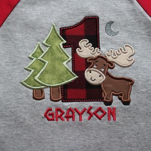 May include: A gray and red raglan shirt with a moose, two pine trees, and a red and black plaid number one. The shirt says "GRAYSON" in red.