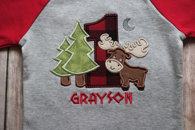 May include: A gray and red raglan shirt with a moose, two pine trees, and a red and black plaid number one. The shirt says "GRAYSON" in red.