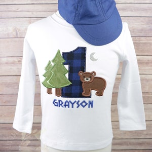 May include: White long-sleeve shirt with a blue and black plaid number one, a green tree, a brown bear, and a silver moon. The shirt is personalized with the name "GRAYSON". A blue baseball cap is on top of the shirt.