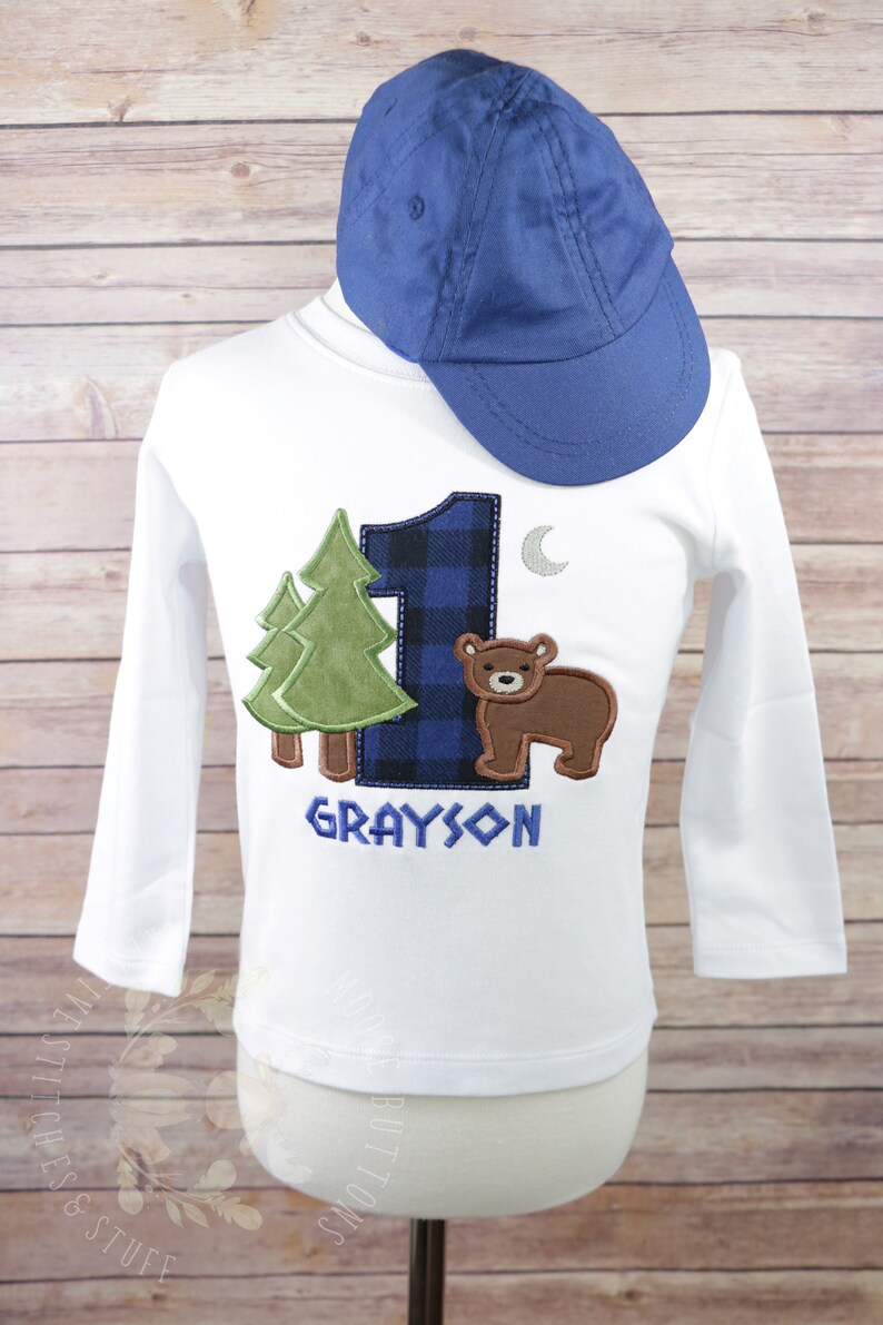 May include: White long-sleeve shirt with a blue and black plaid number one, a green tree, a brown bear, and a silver moon. The shirt is personalized with the name "GRAYSON". A blue baseball cap is on top of the shirt.