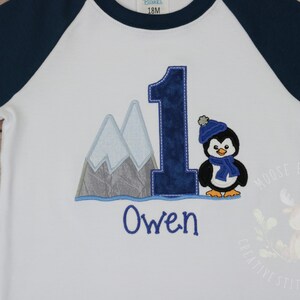 Pu&ograve; includere: Maglietta bianca a maniche lunghe con maniche raglan blu. La maglietta ha un pinguino ricamato con un cappello e una sciarpa blu, un numero uno blu e montagne. Il nome "Owen" &egrave; ricamato sotto il disegno.