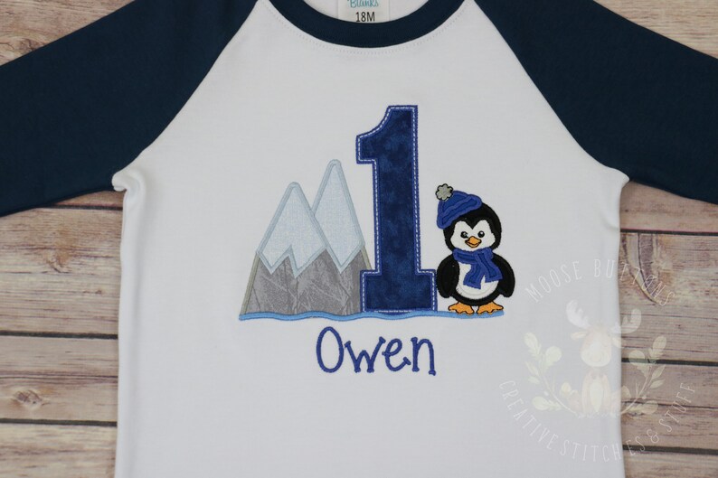Pu&ograve; includere: Maglietta bianca a maniche lunghe con maniche raglan blu. La maglietta ha un pinguino ricamato con un cappello e una sciarpa blu, un numero uno blu e montagne. Il nome "Owen" &egrave; ricamato sotto il disegno.