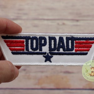 May include: Embroidered patch with the text "TOP DAD" in navy blue, red, and white. The patch features a star and wing design, perfect for adding to clothing or accessories. The patch is rectangular with angled edges.