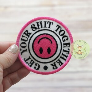 May include: Pink and white embroidered patch with the text "Get Your Shit Together" in a circle around a pink smiley face.