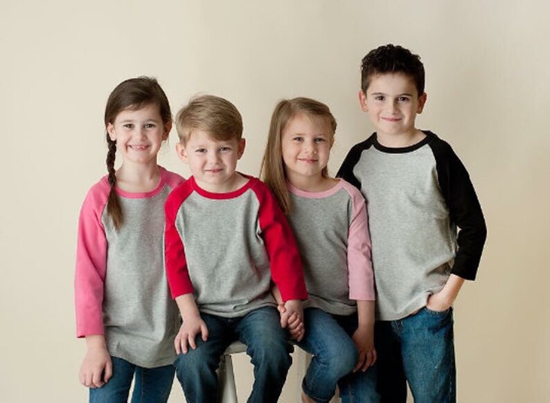 May include: Four children wearing grey and colored baseball shirts with contrasting sleeves. The shirts have a black, pink, red, or black contrasting sleeve. The children are standing in a row, looking at the camera and smiling.