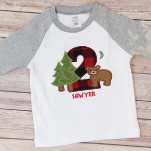 Peut inclure: T-shirt &agrave; manches raglan blanche et grise avec un num&eacute;ro 2 en tissu &eacute;cossais rouge et noir, un arbre vert, un ours brun et le nom "Sawyer" brod&eacute; sur le devant.