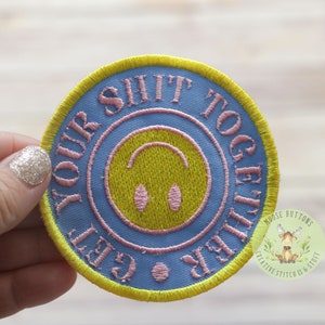 May include: Round embroidered patch with the text "GET YOUR SHIT TOGETHER" in pink lettering on a light blue background. The patch features a yellow smiley face and a yellow border.