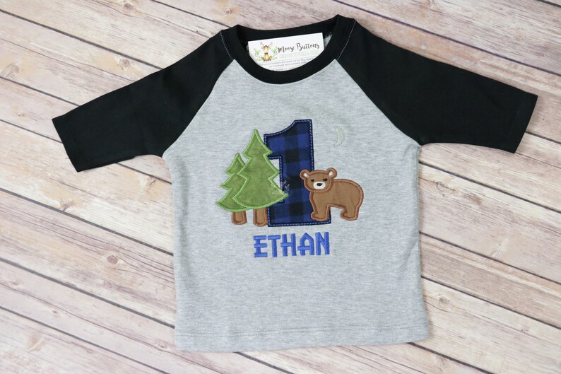 May include: A gray and black baseball tee shirt with a blue and black plaid number one, a green tree, and a brown bear. The name "ETHAN" is embroidered in blue below the number one.