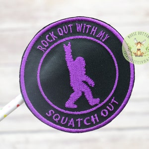 May include: Purple embroidered iron-on patch with a black background. The patch features a silhouette of a Bigfoot with its arm raised in a rock and roll hand gesture. The text "Rock Out With My Squatch Out" is embroidered around the edge of the patch.