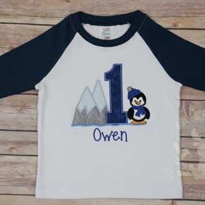 Pu&ograve; includere: Maglietta da baseball bianca e blu navy con un numero uno blu, un pinguino con un cappello e una sciarpa blu, e montagne sullo sfondo. Il nome "Owen" &egrave; ricamato sotto il numero uno.