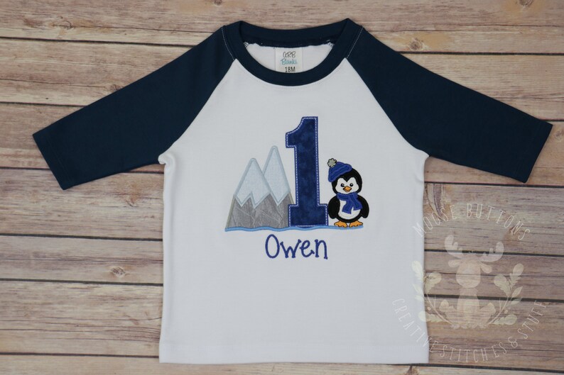 Pu&ograve; includere: Maglietta da baseball bianca e blu navy con un numero uno blu, un pinguino con un cappello e una sciarpa blu, e montagne sullo sfondo. Il nome "Owen" &egrave; ricamato sotto il numero uno.