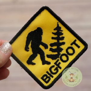 May include: Yellow diamond-shaped warning sign with black border and black text that reads "BIGFOOT". A black silhouette of a Bigfoot walking to the right is in the center of the sign. A small black silhouette of a pine tree is to the right of the Bigfoot.