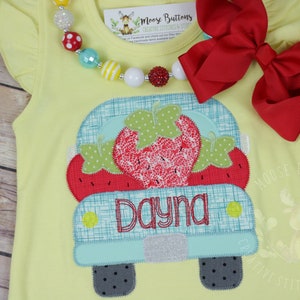 May include: A yellow sleeveless shirt with a red, green, and blue appliqué of a truck carrying a strawberry. The truck has the name "Dayna" embroidered on the back.
