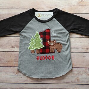 Peut inclure: Un t-shirt de baseball gris et noir avec un num&eacute;ro un en carreaux rouge et noir, deux arbres verts et un ours brun. Le t-shirt dit "HUDSON" sous le motif.