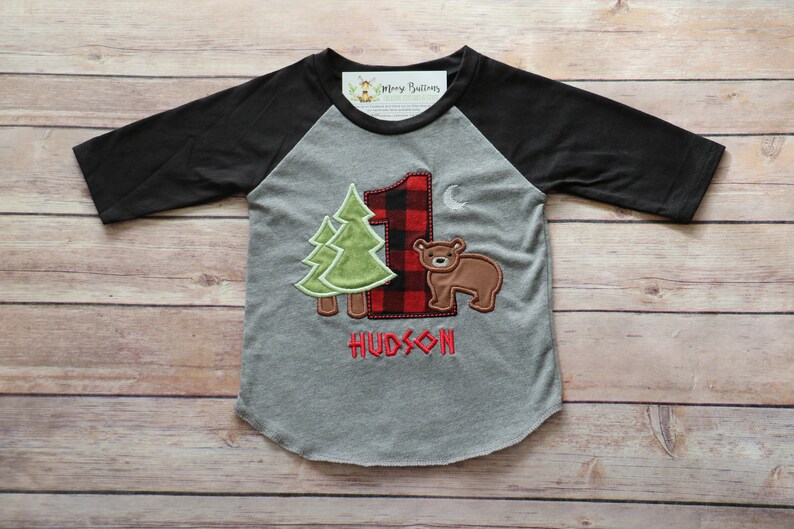 Peut inclure: Un t-shirt de baseball gris et noir avec un num&eacute;ro un en carreaux rouge et noir, deux arbres verts et un ours brun. Le t-shirt dit "HUDSON" sous le motif.