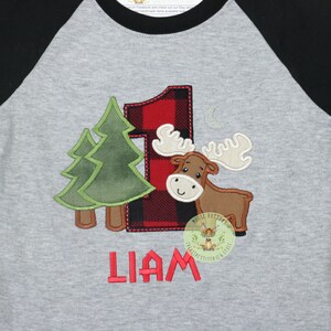 May include: A gray and black raglan t-shirt with a red and black plaid number one, a green tree, a brown moose, and the name Liam embroidered on the front.
