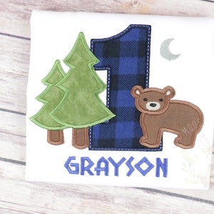 May include: A white onesie with a blue and black plaid number one, two green trees, a brown bear, a silver crescent moon, and the name "GRAYSON" in blue.