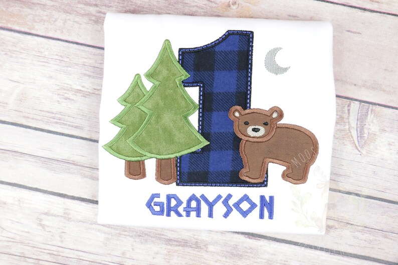 May include: A white onesie with a blue and black plaid number one, two green trees, a brown bear, a silver crescent moon, and the name "GRAYSON" in blue.