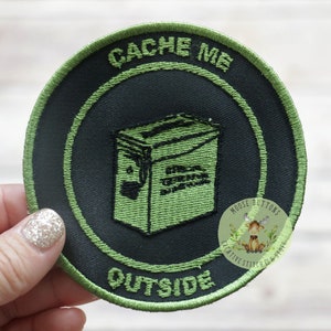 May include: Green and black embroidered patch with the text "CACHE ME OUTSIDE" and a cartoon image of a green metal box. The patch also has a small logo with the text "MOOSE BUTTONS CREATIVE STITCH & STUFFS".