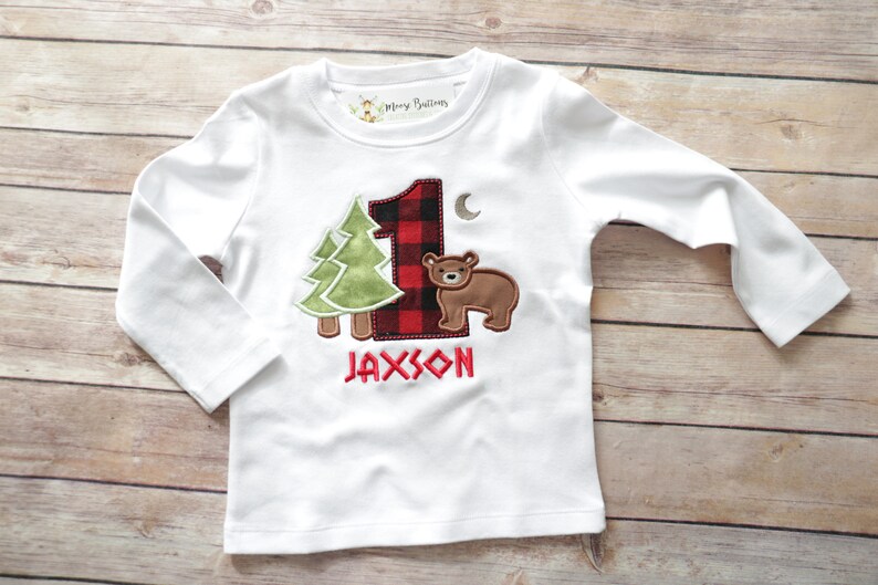 Peut inclure: T-shirt &agrave; manches longues blanc avec un num&eacute;ro un en tissu &eacute;cossais rouge et noir, un arbre vert, un ours brun et le nom Jaxson brod&eacute; sur le devant.