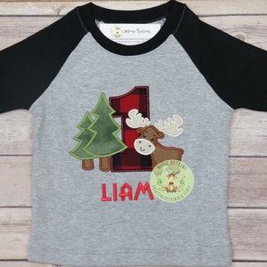 May include: A gray and black baseball tee shirt with a red and black plaid number one, a brown moose, and two green trees. The shirt also has the name "Liam" and a small logo that says "Moose Buttons".
