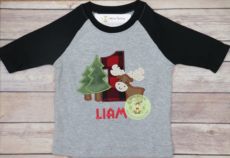 May include: A gray and black baseball tee shirt with a red and black plaid number one, a brown moose, and two green trees. The shirt also has the name "Liam" and a small logo that says "Moose Buttons".