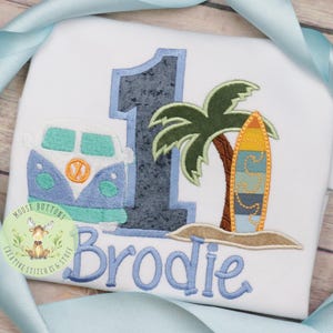 May include: A white onesie with a blue number one, a blue and green VW van, a palm tree, a surfboard, and the name "Brodie" embroidered in blue.