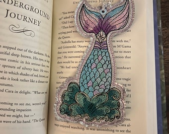 May include: A mermaid tail bookmark with a light blue, pink, and purple design. The tail is embroidered on a white fabric with a green and blue design around the base of the tail. The bookmark is hanging from a silver chain.