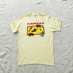 May include: A yellow t-shirt with a red outline of the state of Kansas and the word "Kansas" in red letters. The outline of the state includes images of a buffalo, a combine harvester, a cow, and a city skyline.