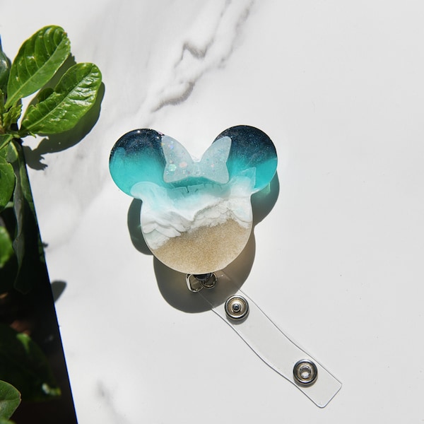 Può includere: Un portabadge in resina blu, bianco e oro a forma di testa di Topolino. Il portabadge è attaccato a un rotolo retrattile per badge in plastica trasparente.