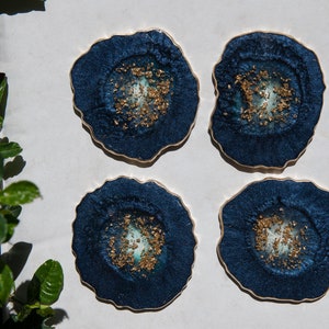 May include: Four blue and gold resin coasters with a geode design. The coasters have a dark blue background with gold flecks and a white centre.