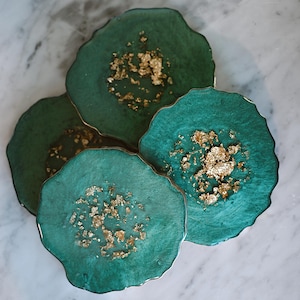 Peut inclure: Quatre dessous de verre en résine turquoise avec des accents de feuilles d'or. Chaque dessous de verre a une forme organique unique et un bord doré. Les dessous de verre sont disposés sur une surface en marbre.