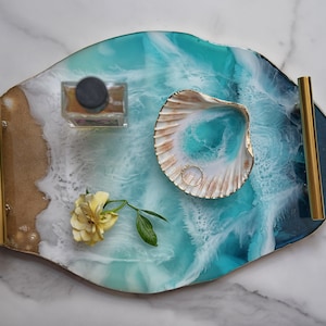 Könnte beinhalten: Ein Tablett aus Harz mit einem blauen und weißen Ozeanwellen-Design, goldenen Akzenten und einer Muschel. Das Tablett hat zwei goldene Griffe an den Seiten.