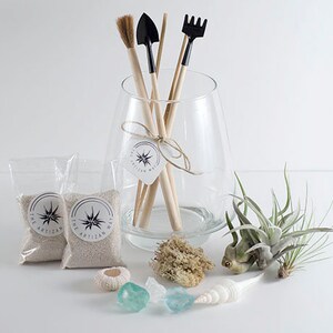 May include: A clear glass vase containing wooden-handled gardening tools, including a brush, shovel, and rake. Two small bags of sand, air plants, a sea urchin shell, two blue glass stones, and a white seashell are arranged in front of the vase.
