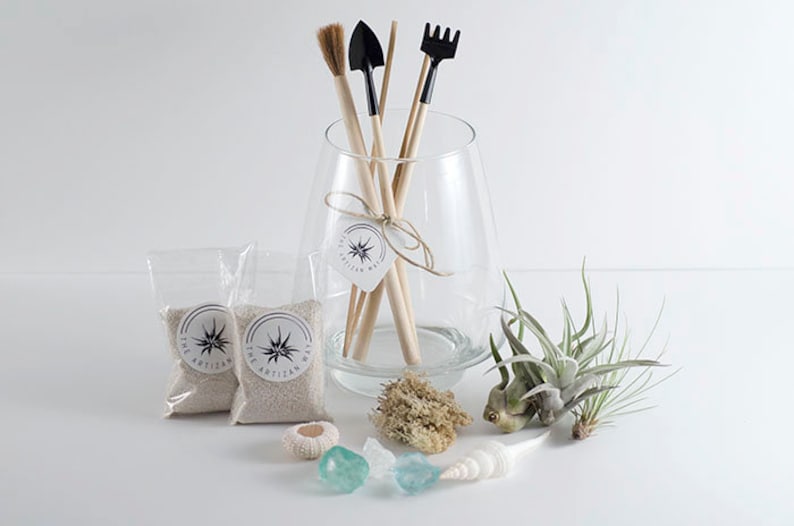 May include: A clear glass vase containing wooden-handled gardening tools, including a brush, shovel, and rake. Two small bags of sand, air plants, a sea urchin shell, two blue glass stones, and a white seashell are arranged in front of the vase.