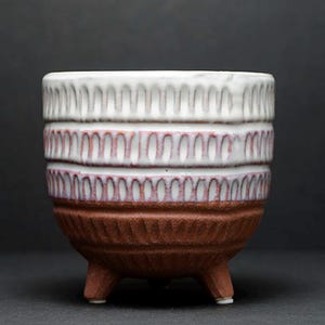 May include: A ceramic planter with three short legs, featuring a textured terracotta base and a white glazed upper section. The exterior is decorated with repeating vertical carved grooves that create a rhythmic pattern around the entire vessel.